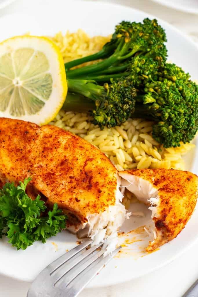 Close up view of a fork holding a bite of seasoned halibut with rice and broccoli.
