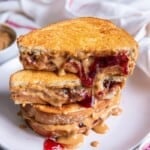 A stack of peanut butter and jelly sandwiches on a white plate, the top sandwich is cut in half.