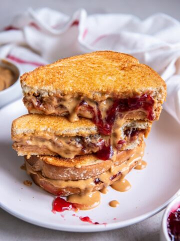 A stack of peanut butter and jelly sandwiches on a white plate, the top sandwich is cut in half.