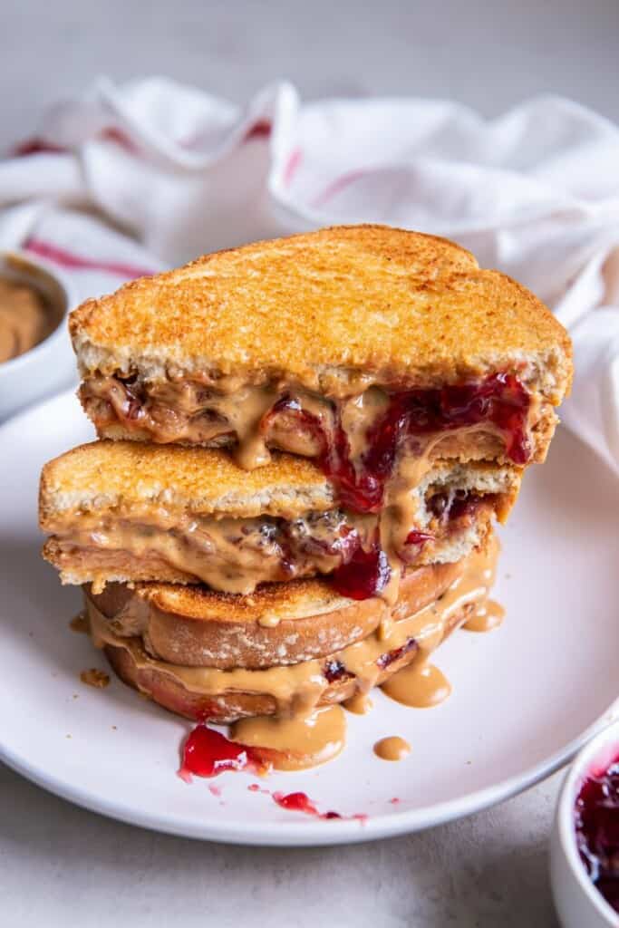 A stack of peanut butter and jelly sandwiches on a white plate, the top sandwich is cut in half.