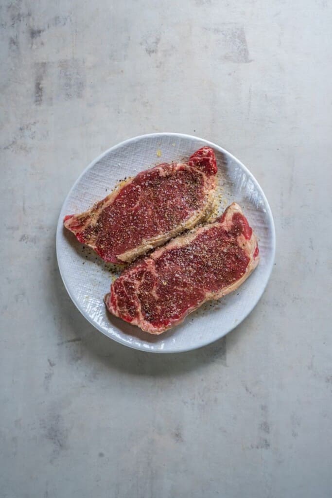 Two seasoned new york strip steaks on a white plate.