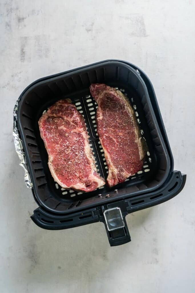 Two seasoned new york strip steaks resting in a black air fryer basket.