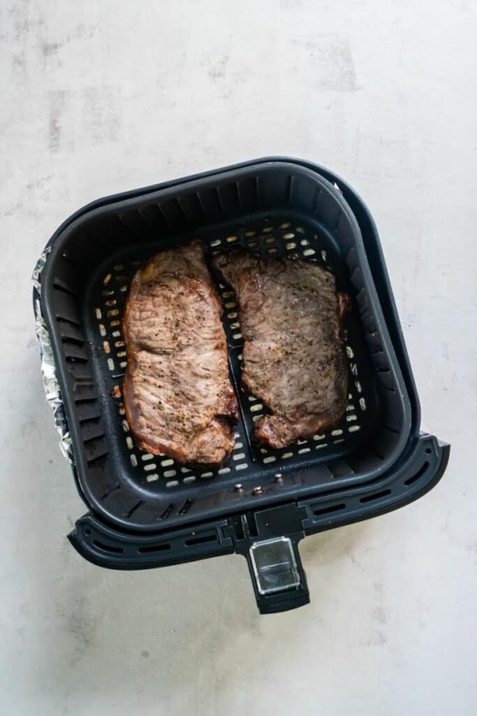 Two prepared new york strip steaks resting in a black air fryer basket.