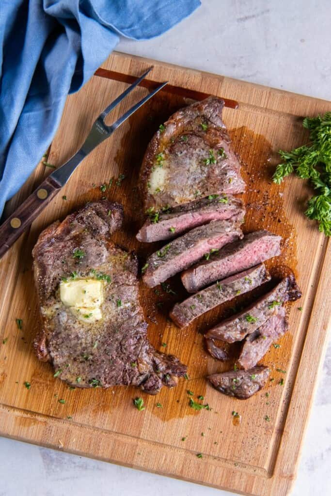 A New York Strip Steak with a pat of butter next to a second sliced steak on a wooden cutting board.