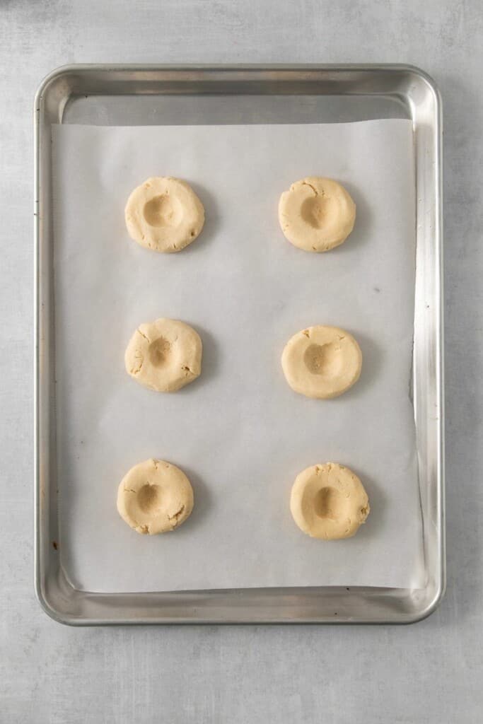 Six cookie dough balls flattened with a thumbprint in the middle on a parchment lined baking sheet.