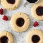 Six raspberry thumbprint cookies surrounded by raspberries.