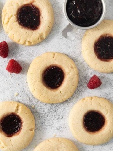 Six raspberry thumbprint cookies surrounded by raspberries.