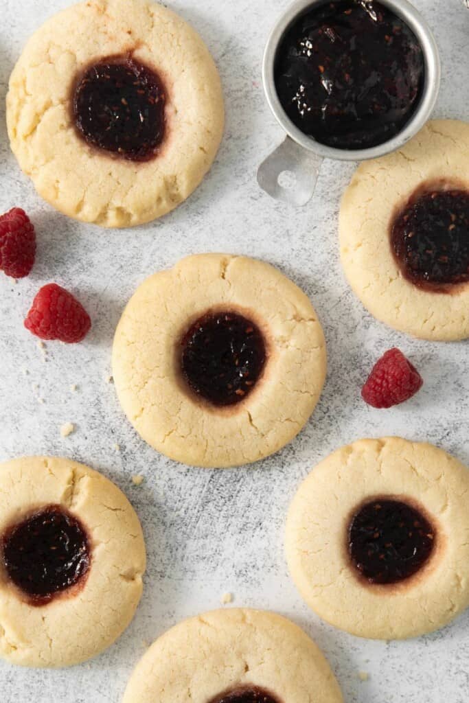 Six raspberry thumbprint cookies surrounded by raspberries.