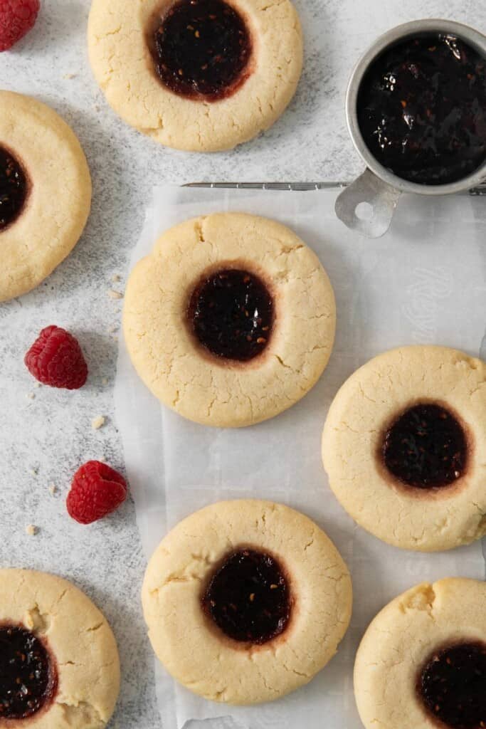 A handful of baked raspberry thumbprint cookies with a few raspberries on parchment paper.