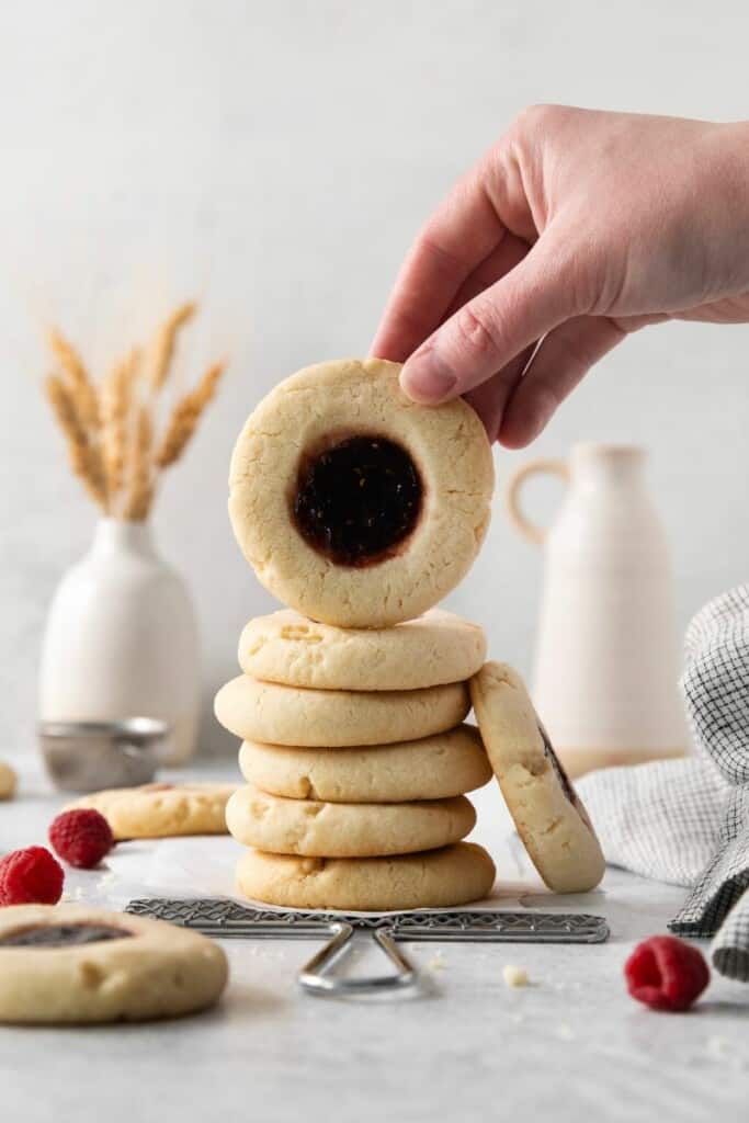 Stack of raspberry thumbprint cookies. Top cookie lifted by hand to show the top.