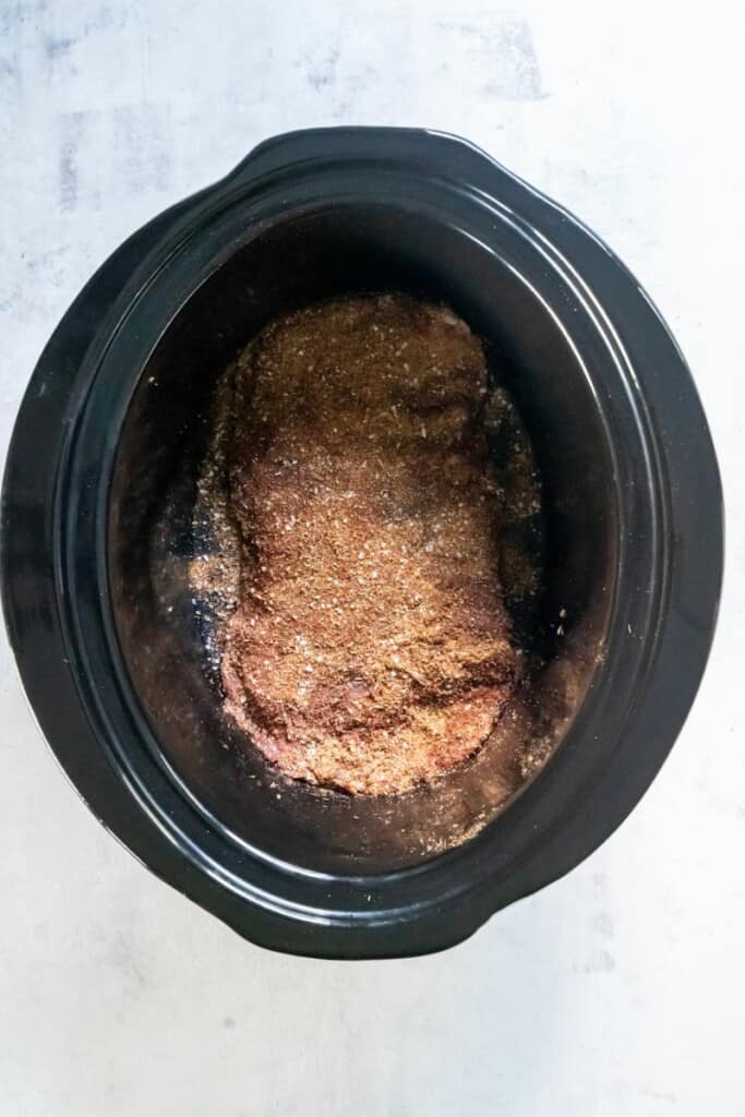 Seasonings and spices sprinkled on a skirt steak in a black crock pot.