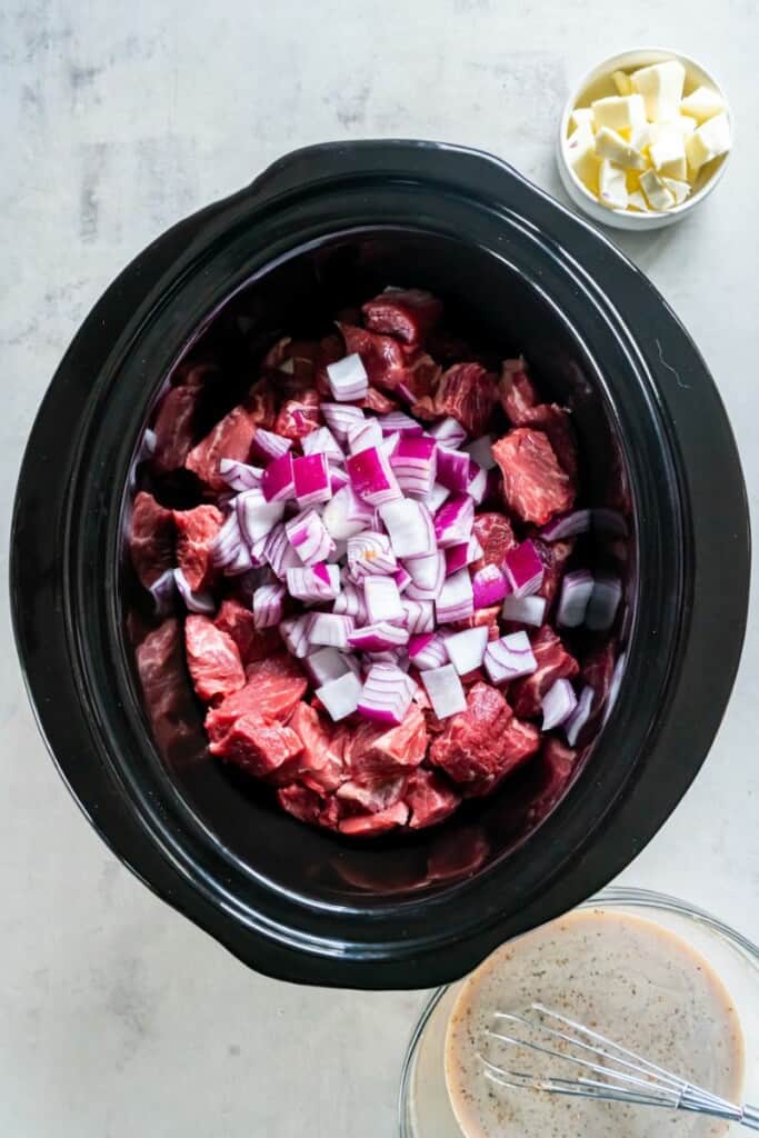 Cubes of steak with spices and seasonings in a black crock pot.