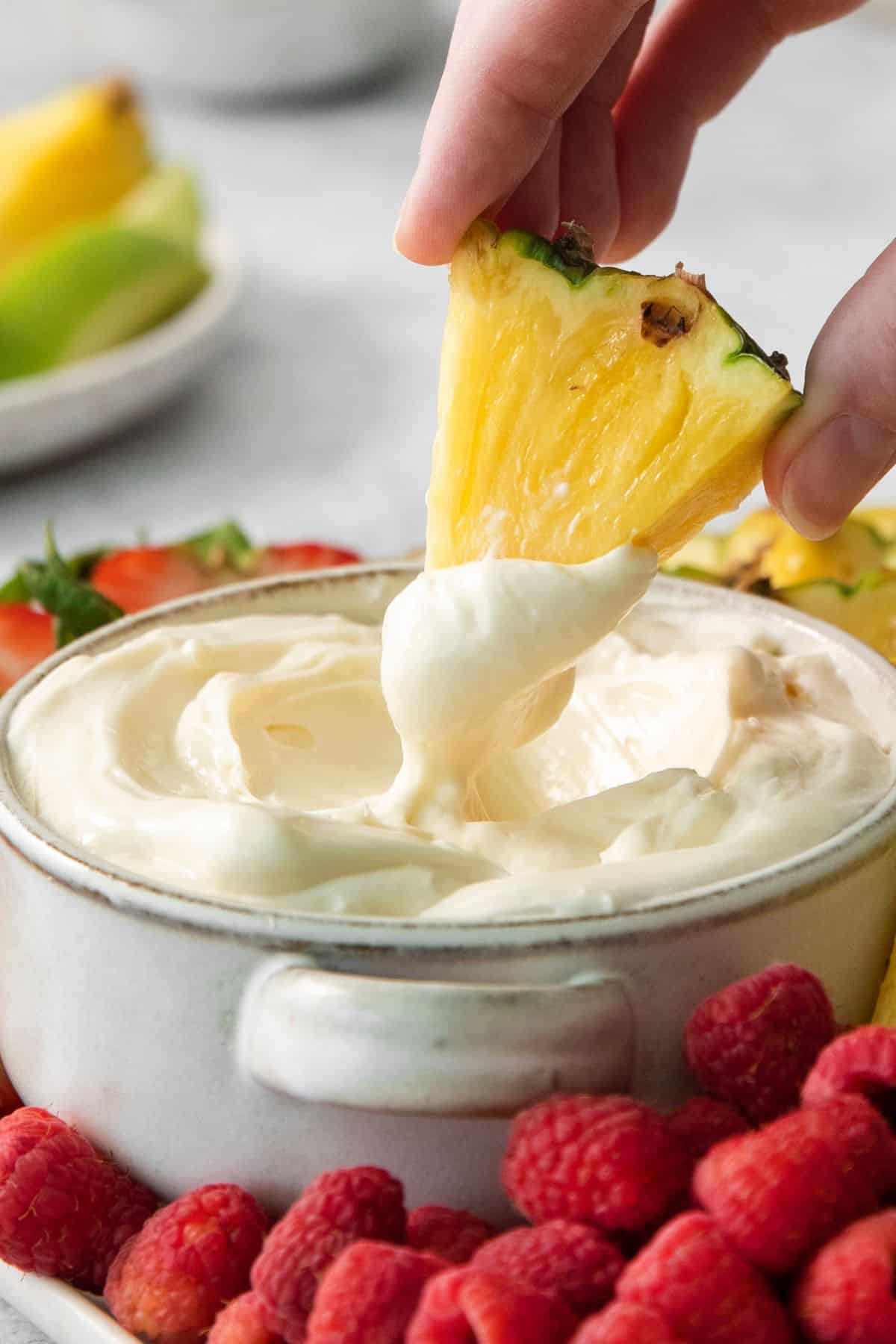 Dipping a piece of pineapple into the fruit dip recipe with marshmallow dip to show how easy it is for dipping.