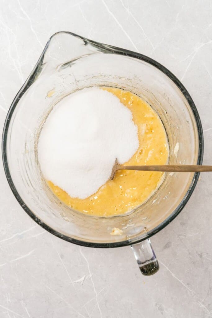 Adding sugar to banana mixture for banana bread in a mixing bowl.