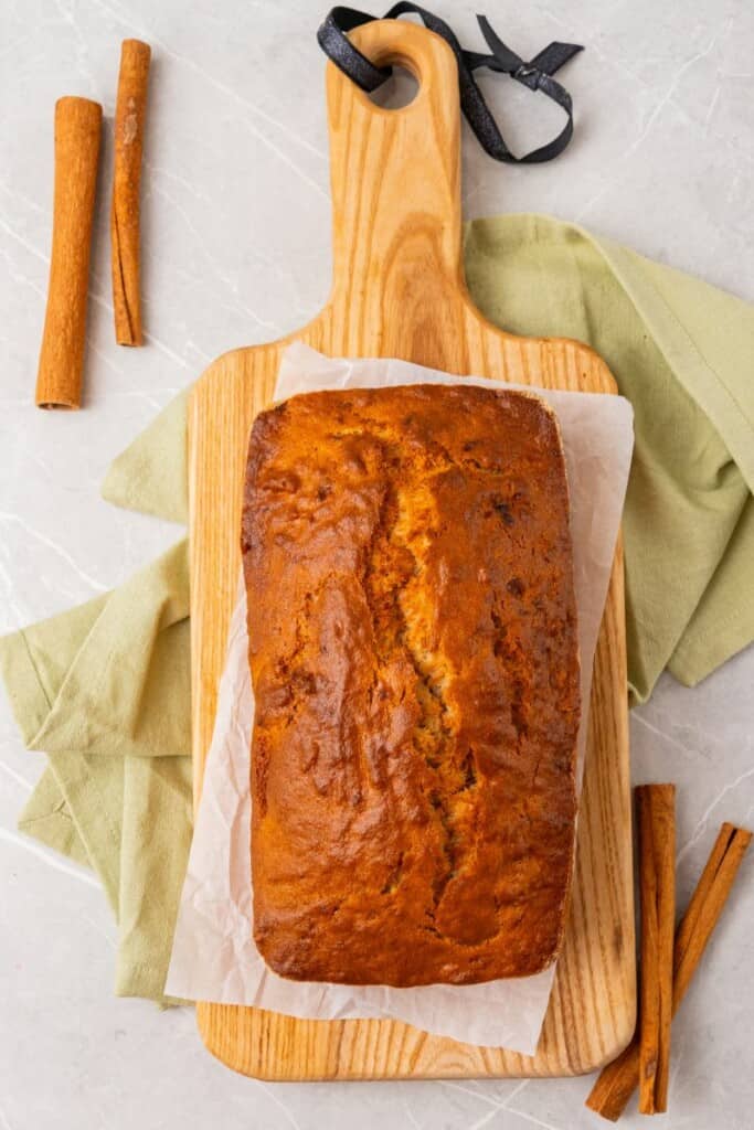 A loaf of banana bread on parchment paper on a wooden cutting board with cinnamon sticks in the background.
