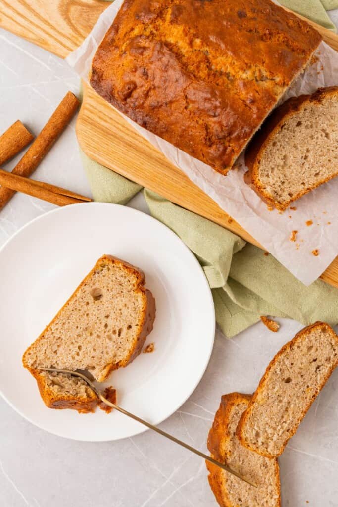 A fork cutting into a slice of banana bread on a saucer. Additional slices and the remaining loaf in the background.