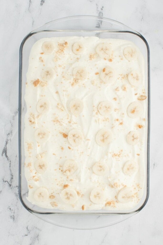 adding fresh banana slices and cookie crumbs on top of homemade banana pudding recipe.