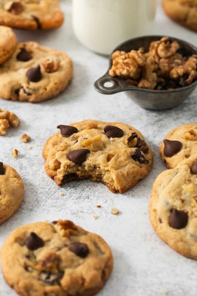 Several baked walnut chocolate chip cookies, the center cookie has a bite removed, a portion of walnuts and a glass of milk.