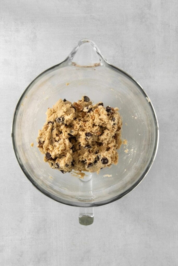 Adding chocolate chips and walnuts to cookie dough base in a clear mixing bowl.
