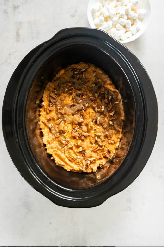 Adding brown sugar and pecans to mashed sweet potatoes in a black crock pot.