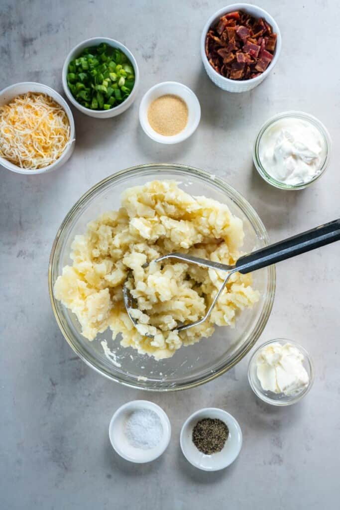 Potato flesh in a clear mixing bowl with butter being mashed with a potato masher.