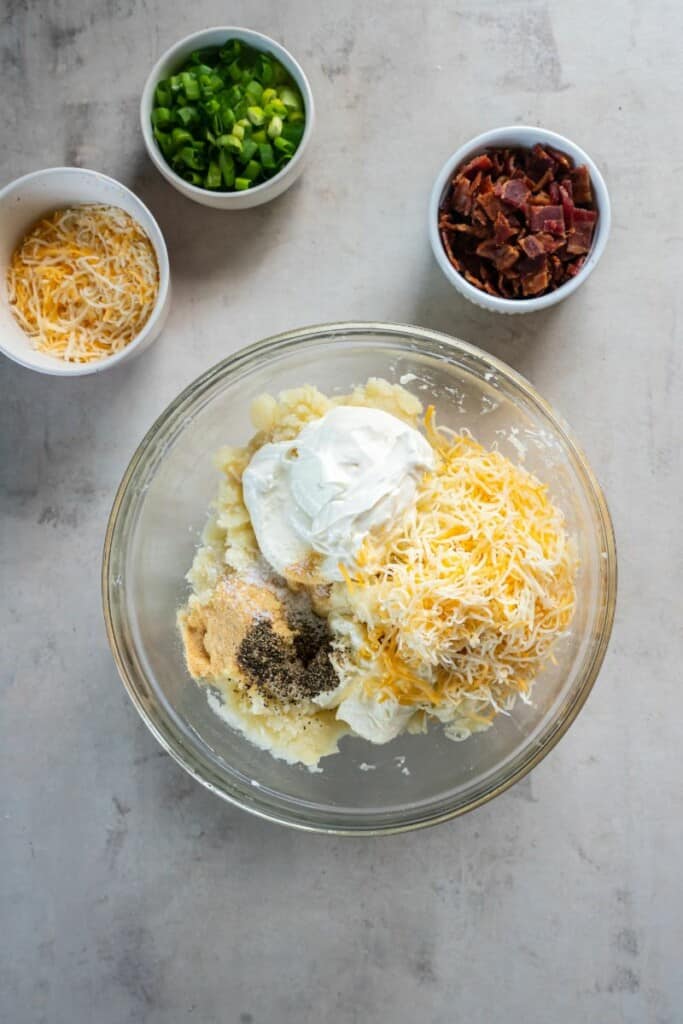 Adding cheddar cheese, sour cream, cream cheese and seasonings to mashed potatoes in a clear mixing bowl.