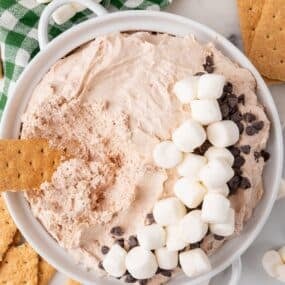 Hot cocoa dip topped with marshmallows and chocolate chips with a graham cracker dipped in it.