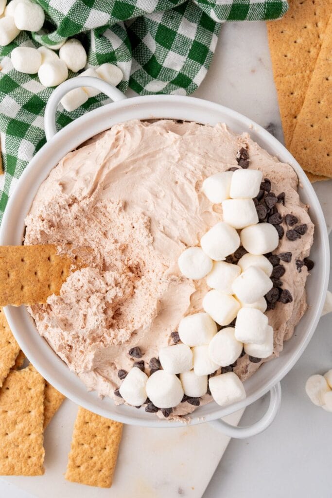 Hot cocoa dip topped with marshmallows and chocolate chips with a graham cracker dipped in it.
