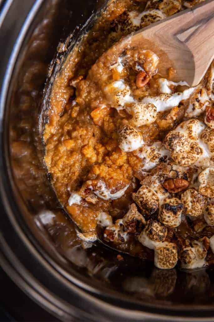Close up of crock pot sweet potato casserole with marshmallows on top and a wooden spoon inside the crock pot with it.