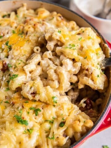 Dutch oven mac and cheese in a dutch oven pot being scooped up by a serving spoon so you can see the cheesiness and bacon.