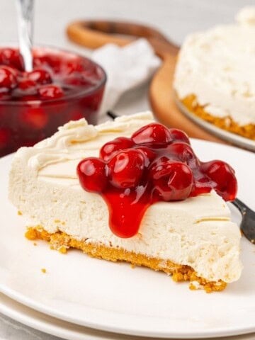 A slice of no bake cheesecake with Cool Whip on a white plate with cherry pie filling on top spilling off the sides.