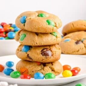 Peanut butter M&M cookies stacked on top of each other on a white plate with peanut butter M&Ms around it.
