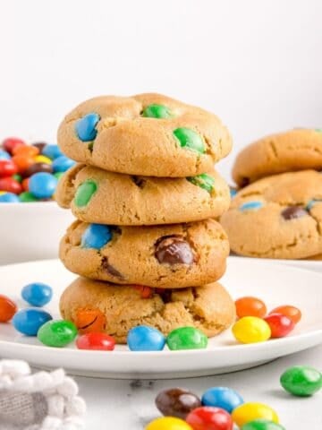 Peanut butter M&M cookies stacked on top of each other on a white plate with peanut butter M&Ms around it.