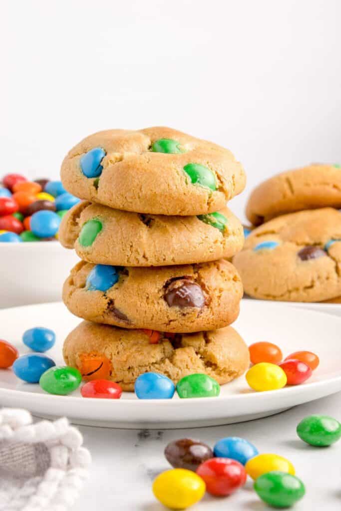 Peanut butter M&M cookies stacked on top of each other on a white plate with peanut butter M&Ms around it.