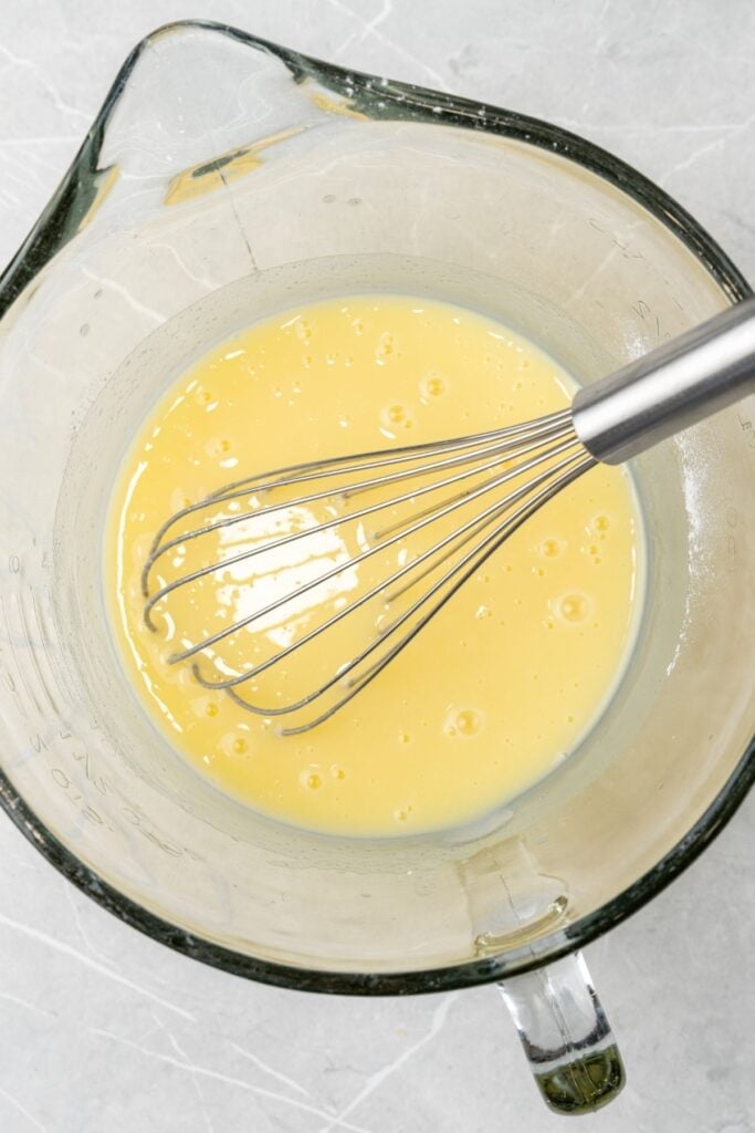 Vanilla pudding mixed with sugar and milk in a mixing bowl with a whisk inside.