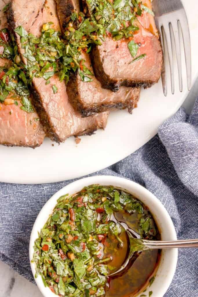 Homemade chimichurri in a bowl with sliced flank steak on a plate. 