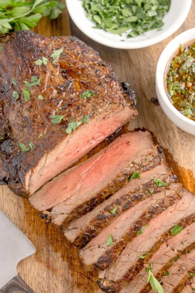 Sliced london broil beef sprinkled with herbs on cutting board. 