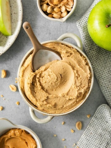 Bowl of peanut butter dip with green apples and peanuts surrounding.