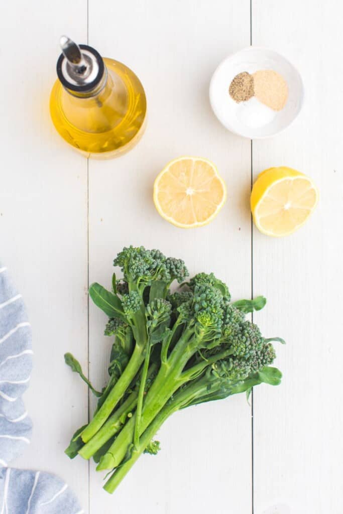 bunch of baby broccoli next to a halved lemon, seasonings and olive oil