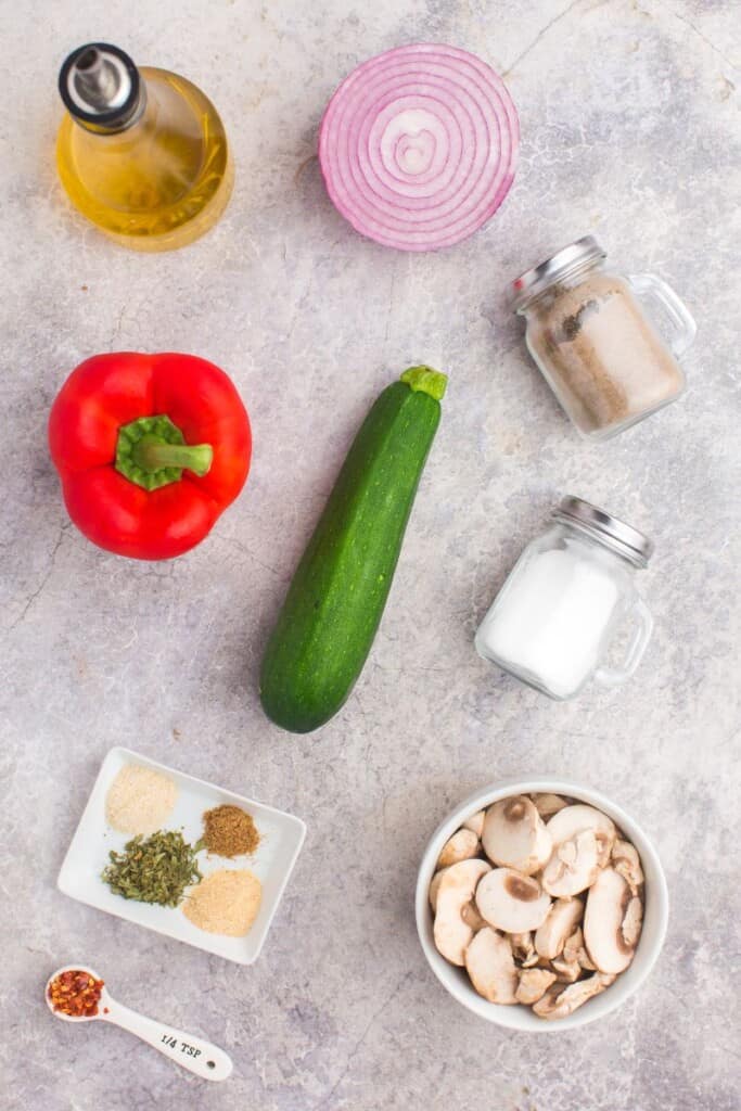 a zucchini, red bell pepper, red onion half, mushrooms, olive oil, and spices on a counter