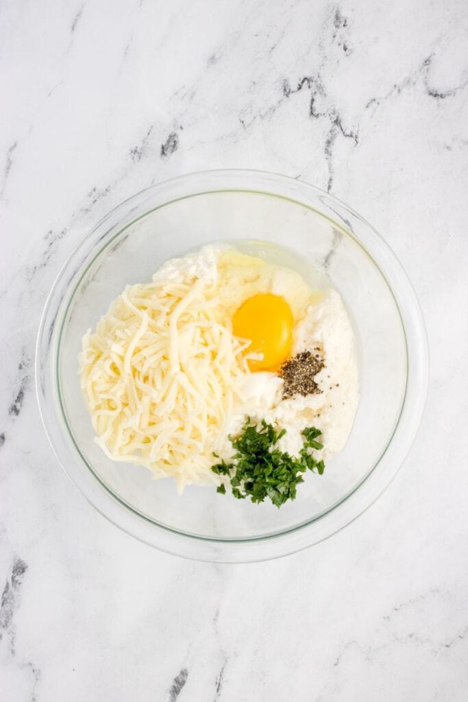 mixing ricotta, eggs, spices, shredded cheese and parsley in a glass bowl.