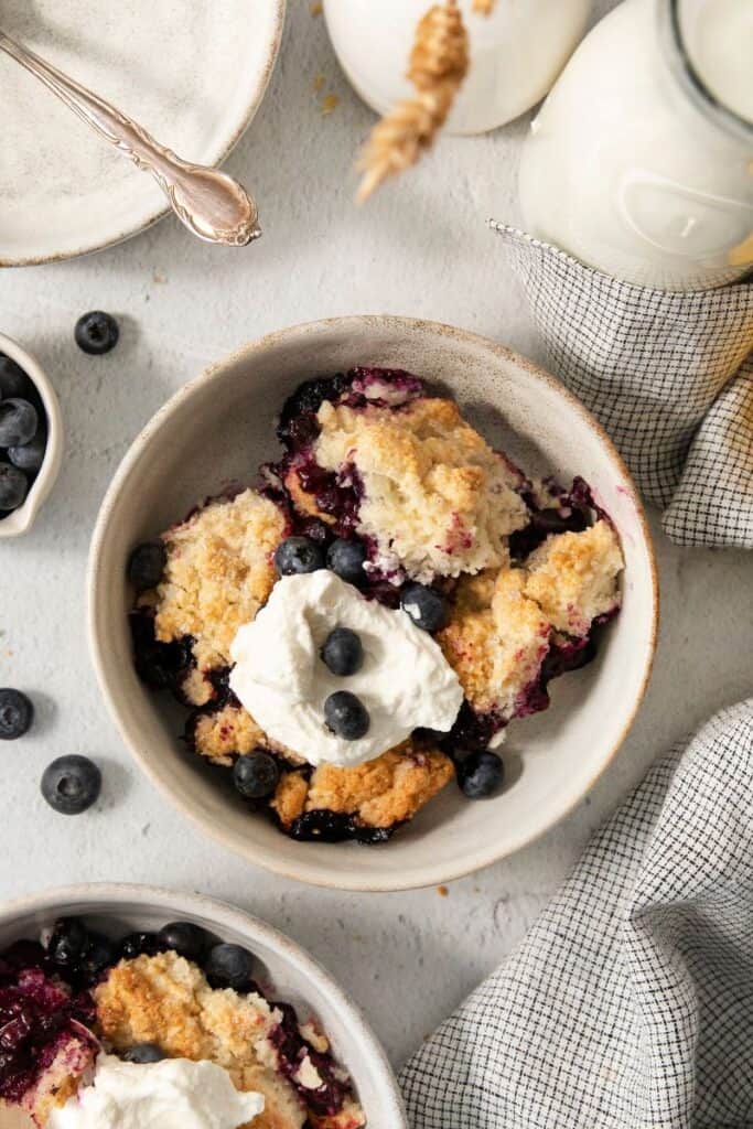 overhead shot of blueberry cobbler served in a bowl with whipped topping and fresh blueberries