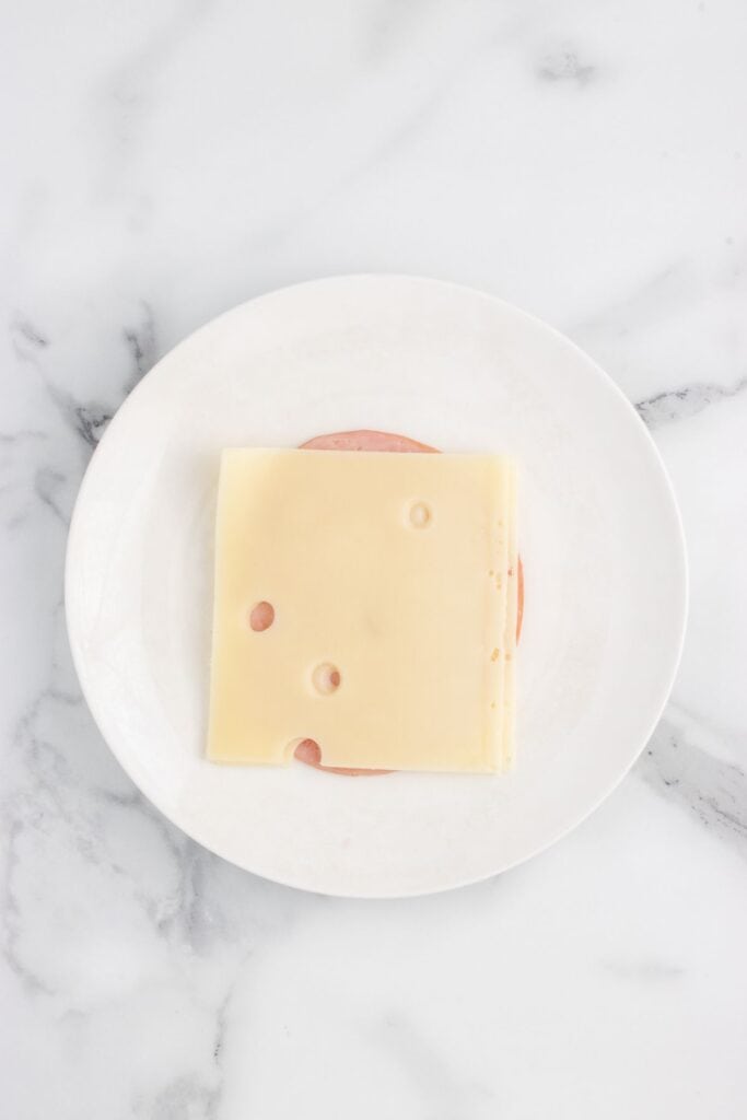 placing a slice of ham onto two pieces of sliced swiss cheese on a plate. 