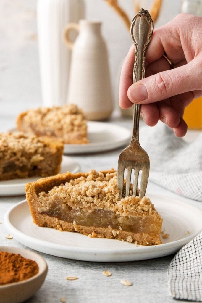 Apple pie with graham cracker crust slice on a white plate with a fork about to take a piece off.