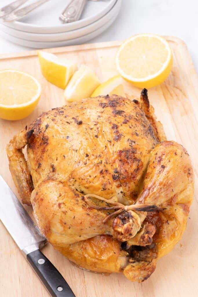 placing oven roasted whole chicken on a wooden cutting board with sliced lemon halves