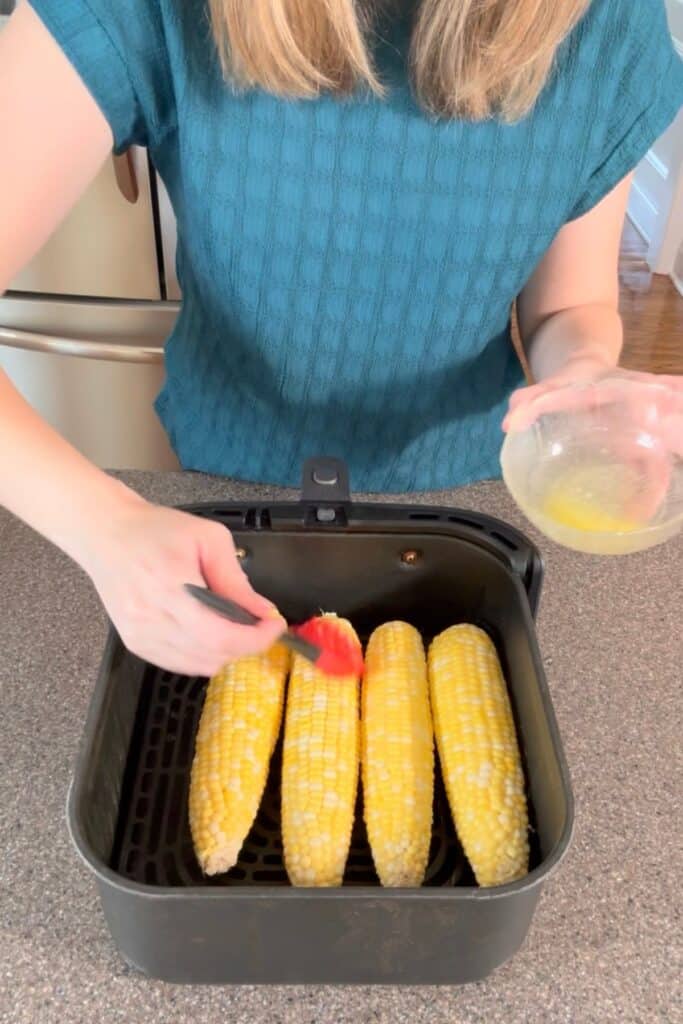 A woman basting corn on the cob sitting in the air fryer with butter and salt.