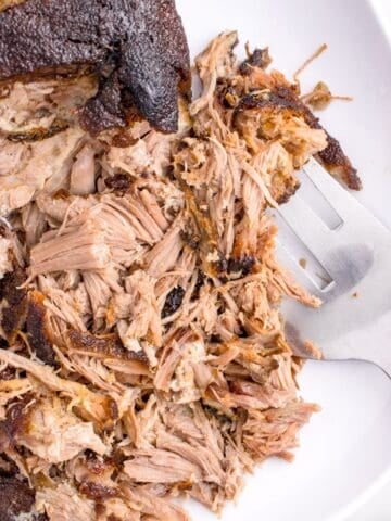 Oven roasted pulled pork cooked and being shredded with a fork on a plate.