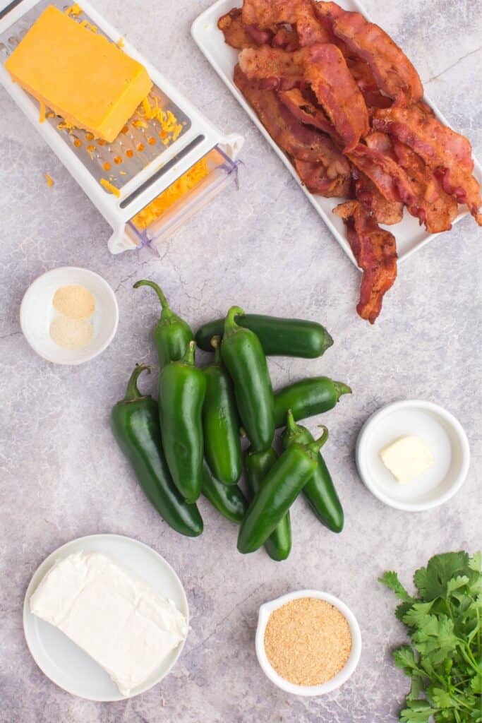 Ingredients needed to make jalapeno poppers in the oven: jalapeno peppers, cream cheese, garlic powder, onion powder, cheddar cheese, breadcrumbs, butter, bacon, and cilantro.