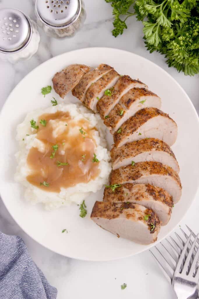 Cooked pork tenderloin sliced and served on a white plate with mashed potatoes and gravy.