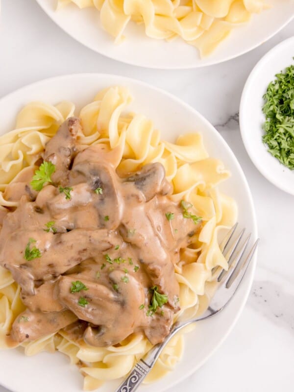 Beef stroganoff served over a bed of egg noodles on a white plate with a fork. Parsley garnish on top.